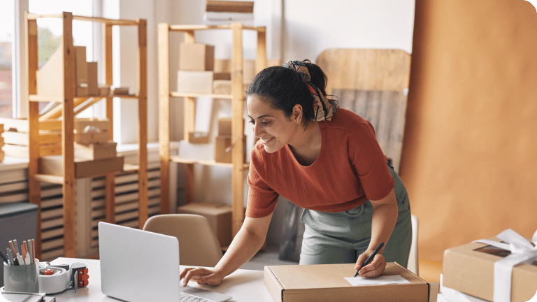 A small business owner leans over their desk, looking at their laptop while writing something on a package label.