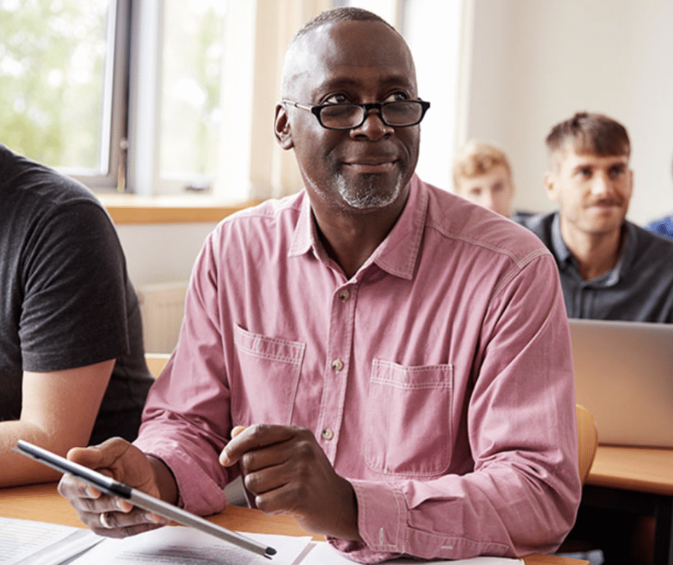 Classroom of people sit looking ahead with one holding a tablet.