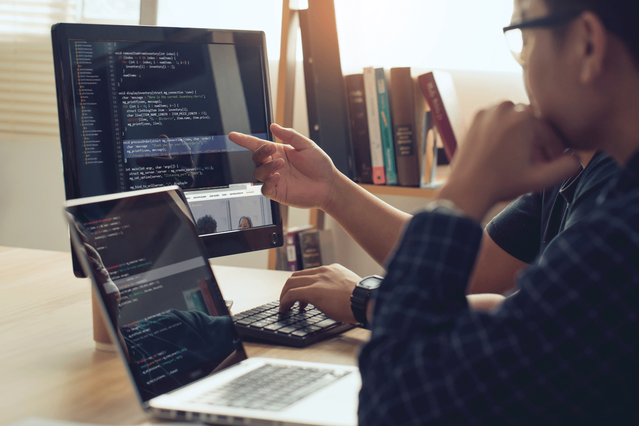 An open laptop and computer monitor with website developer code are looked at by two people sitting at a desk.