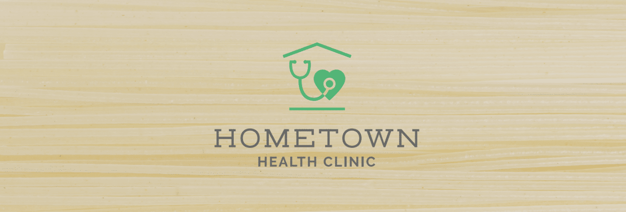 Hometown Health Clinic in ash grey font on a wooden background and a green hut with a heart and stethoscope