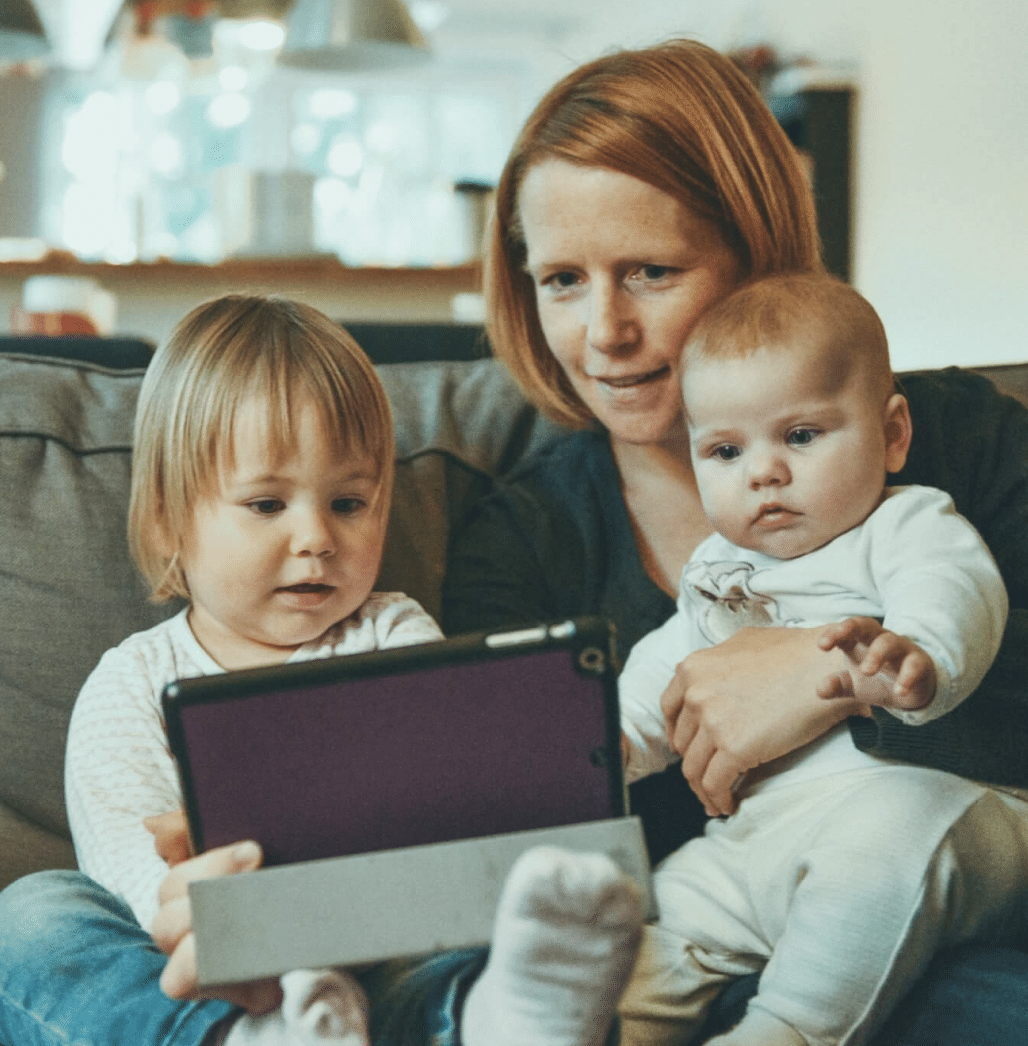 A person sits with a baby on her lap and toddler next to them with an iPad across the toddlers legs.