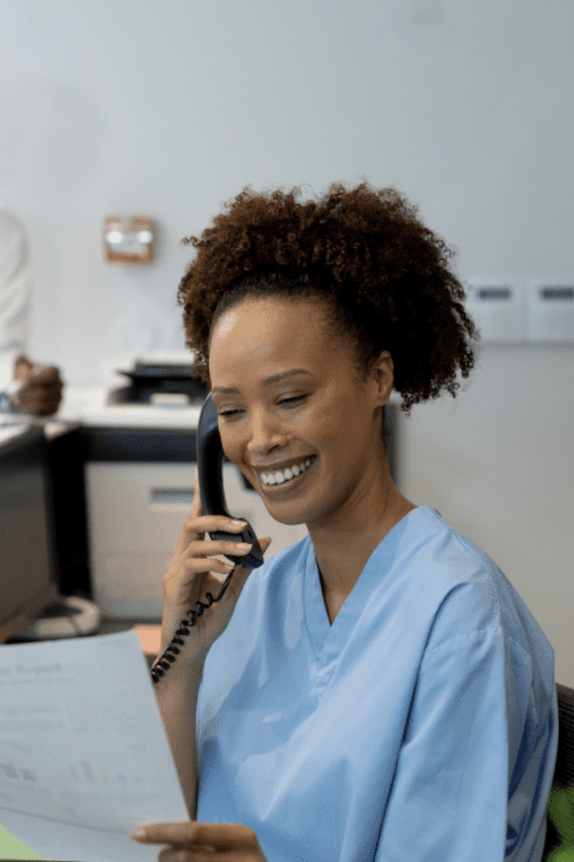 A nurse sits at a desk answering a corded phone, holding a piece of paper in the other hand.