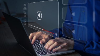 Hands hover over a laptop keyboard with a holographic image of a media player with the play icon displayed.
