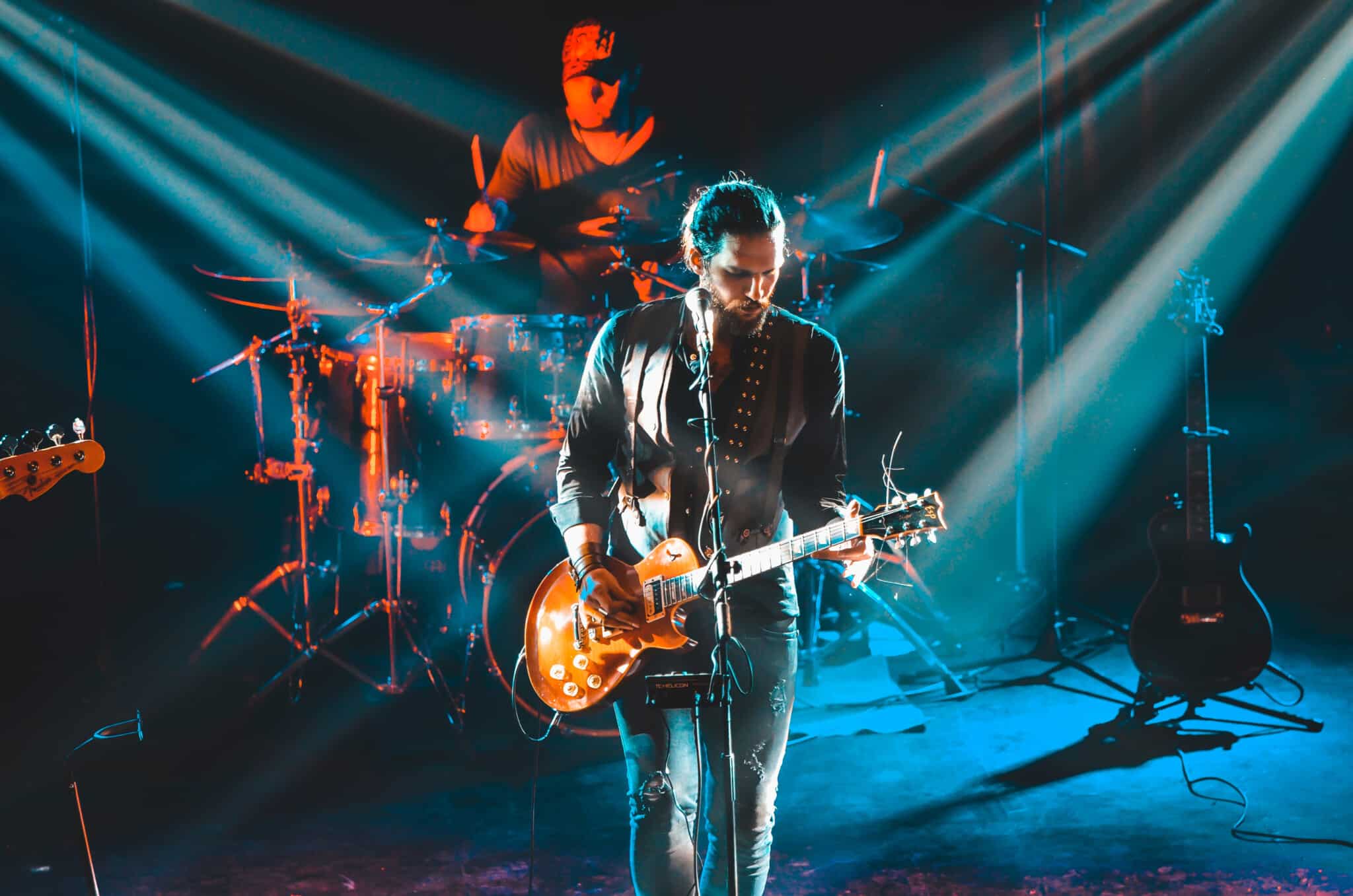 A band performs on stage with a drummer in the background and guitar player in front of a microphone.