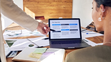 A person sits in front of an open laptop, with papers and notes laying about a table, with another persons arm point to linking strategy on the laptop.