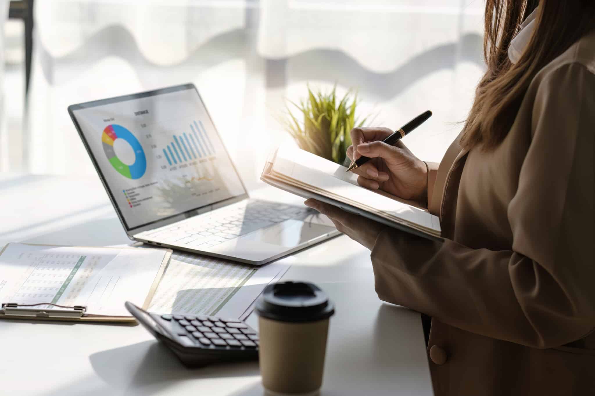 Person reviews data charts and other metrics on laptop while writing notes for monthly budgeting.