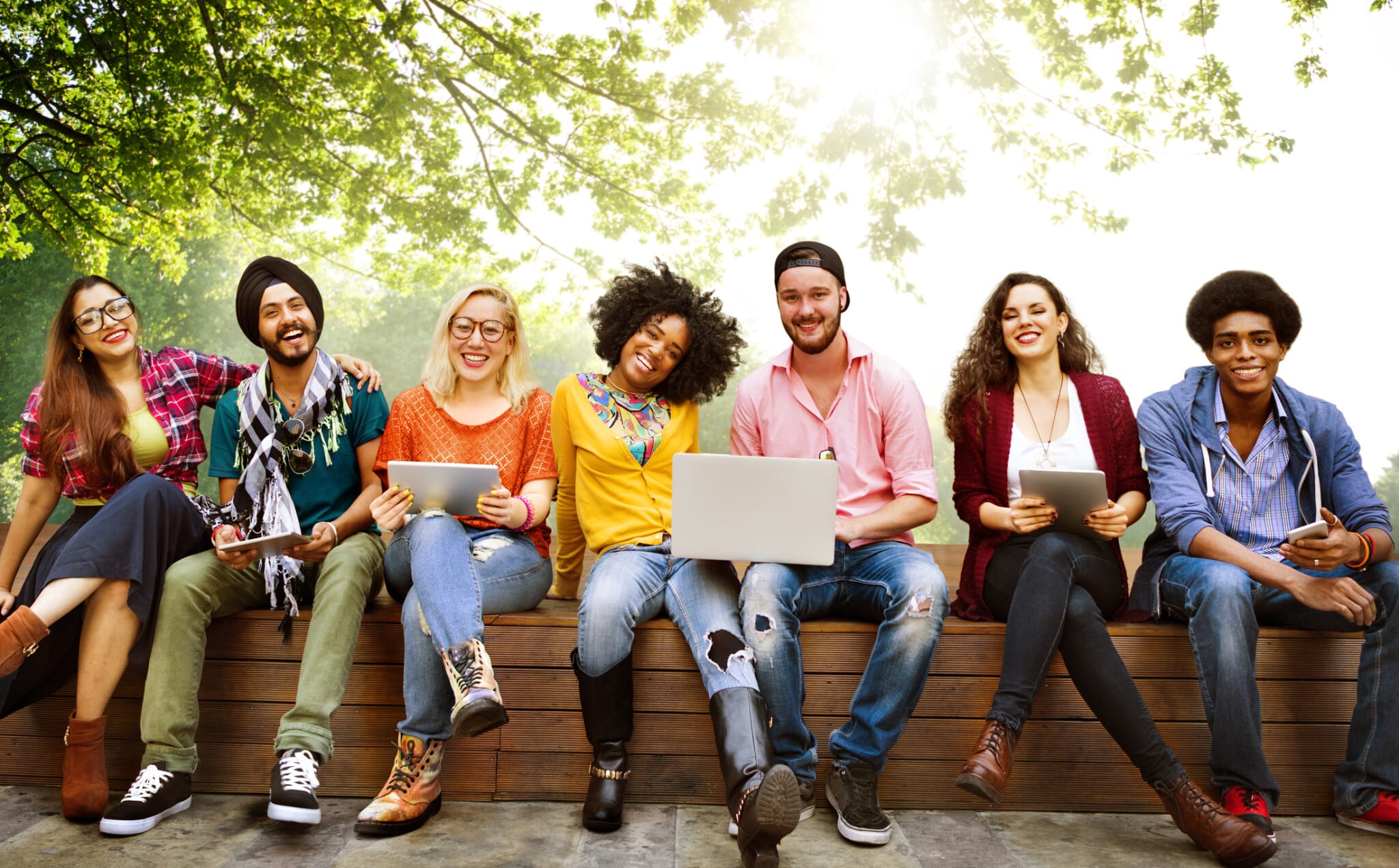 Students sit outside on a bench with various electronics out for studying.
