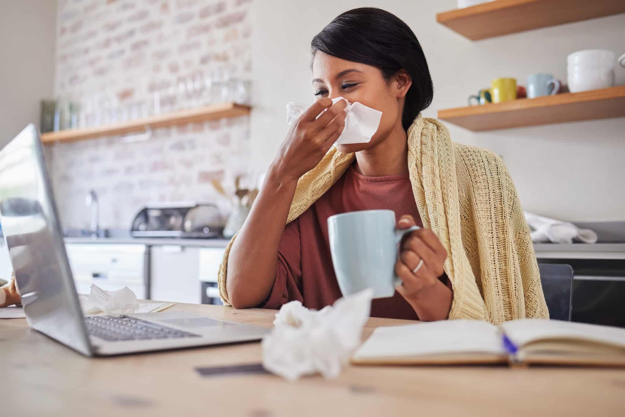 Sick patient sit in front of laptop with tissues at their nose, tea in their hand, for their virtual doctor appointment.