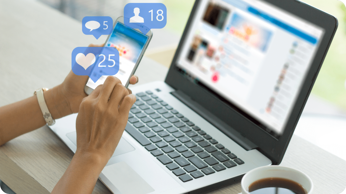 An open laptop sits open in the background of a mobile phone with holographic comment and like bubble icons over it.