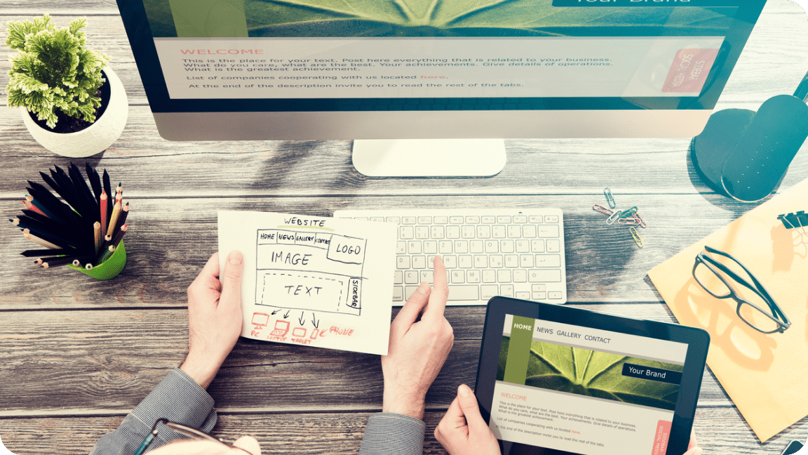 One person holds a design mockup and the other a tablet as they sit in front of a computer screen.