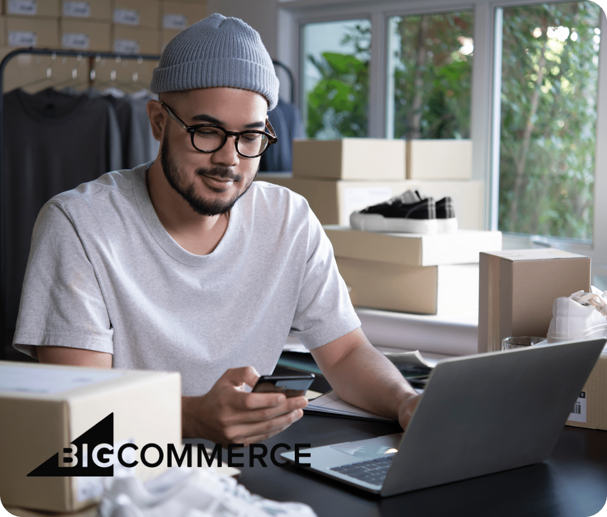 Person sits at their desk with their laptop open, and cellphone in hand, packages on their desk preparing to ship.