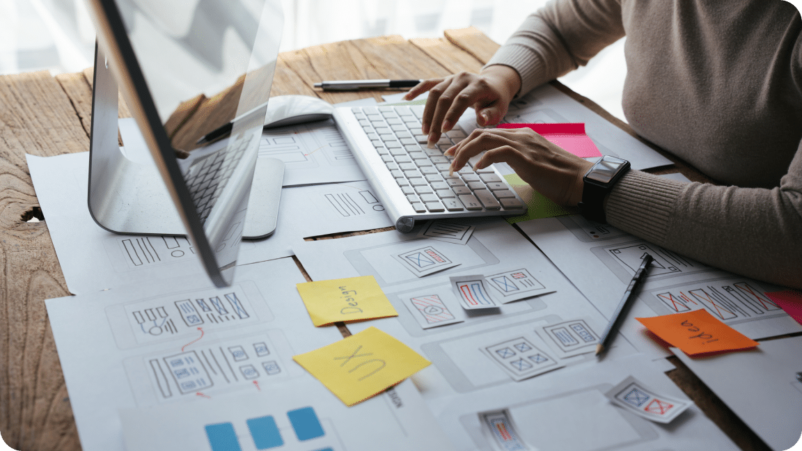 A lady typing on a wireless keyboard with the monitor and papers with sticky notes having touchup points written depicting Brand values.