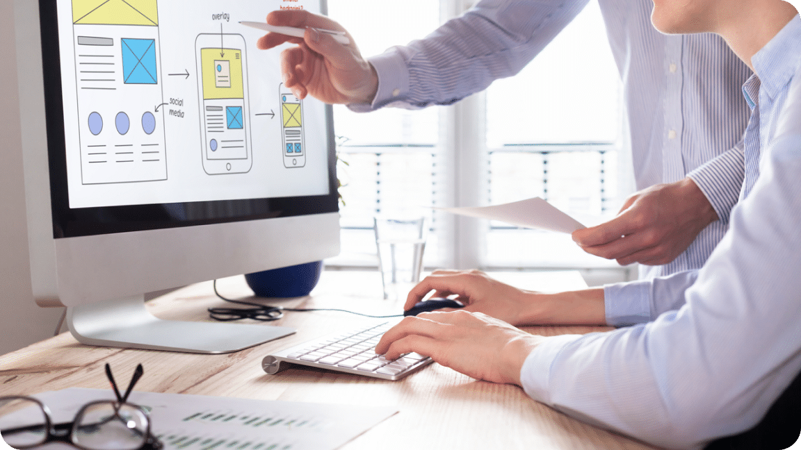 Design layout on a computer monitor is reviewed, with papers in hand and edits made.