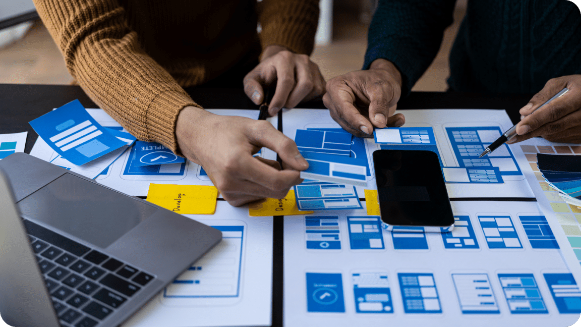 Two persons discussing the website designs with a laptop, notes, and website structures on paper on a table, showing custom CRM development.