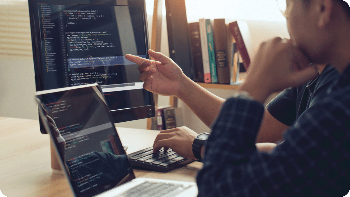 Two people look at a computer screen with a laptop to the side of it looking at website code.
