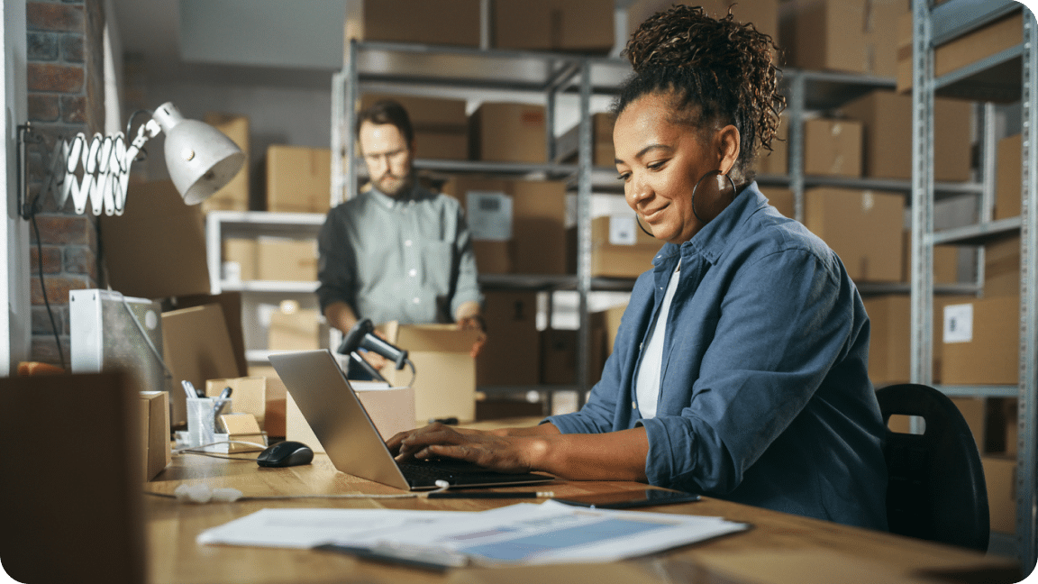 One person looks at a laptop while another packages a shipping box.