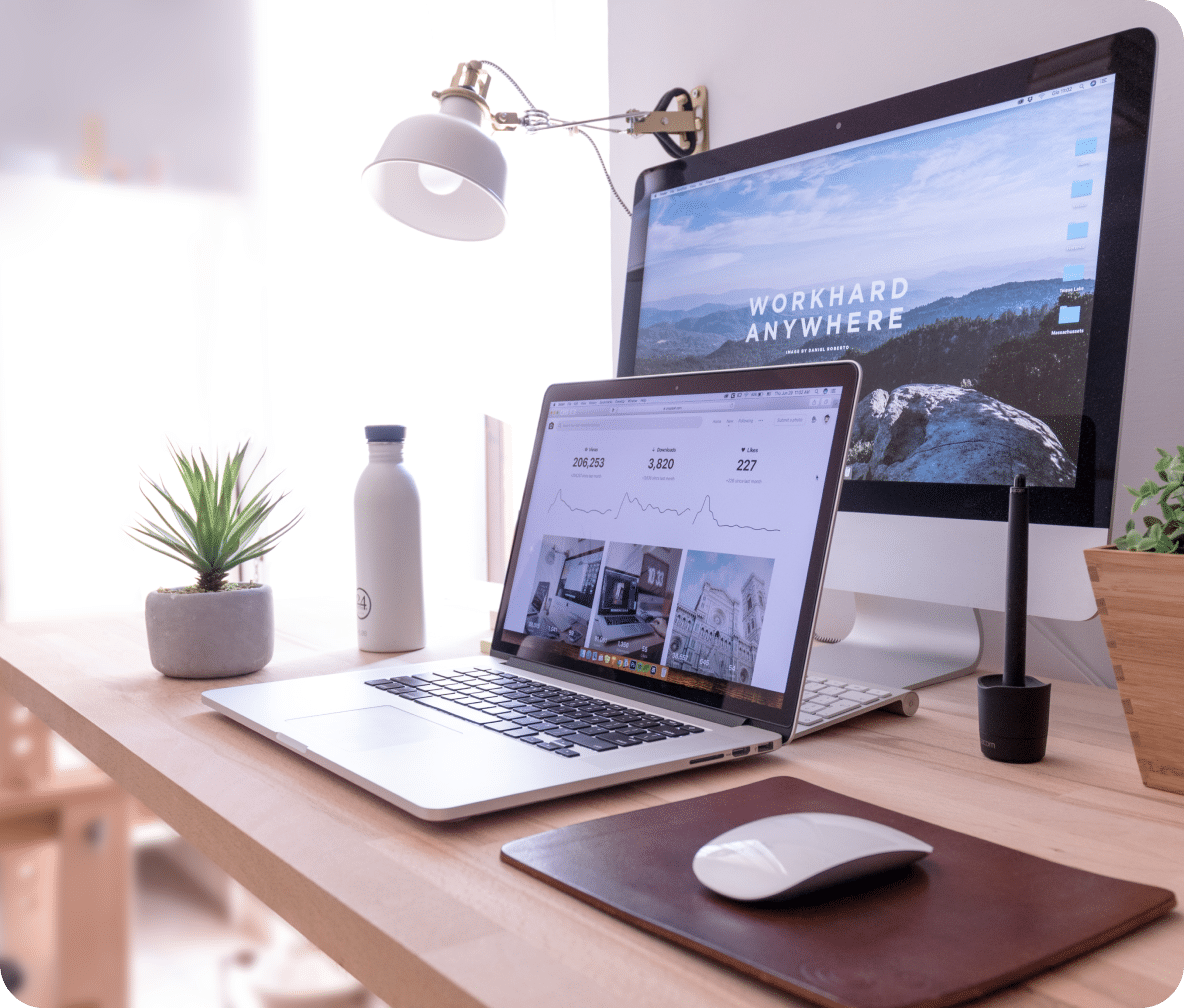Laptop sits in front of a large monitor with a mouse to the side on a desk.