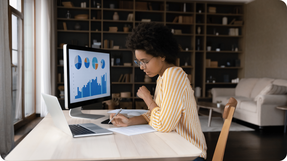 Data charts and graphs show on a monitor as a person sitting at a desk makes notes.