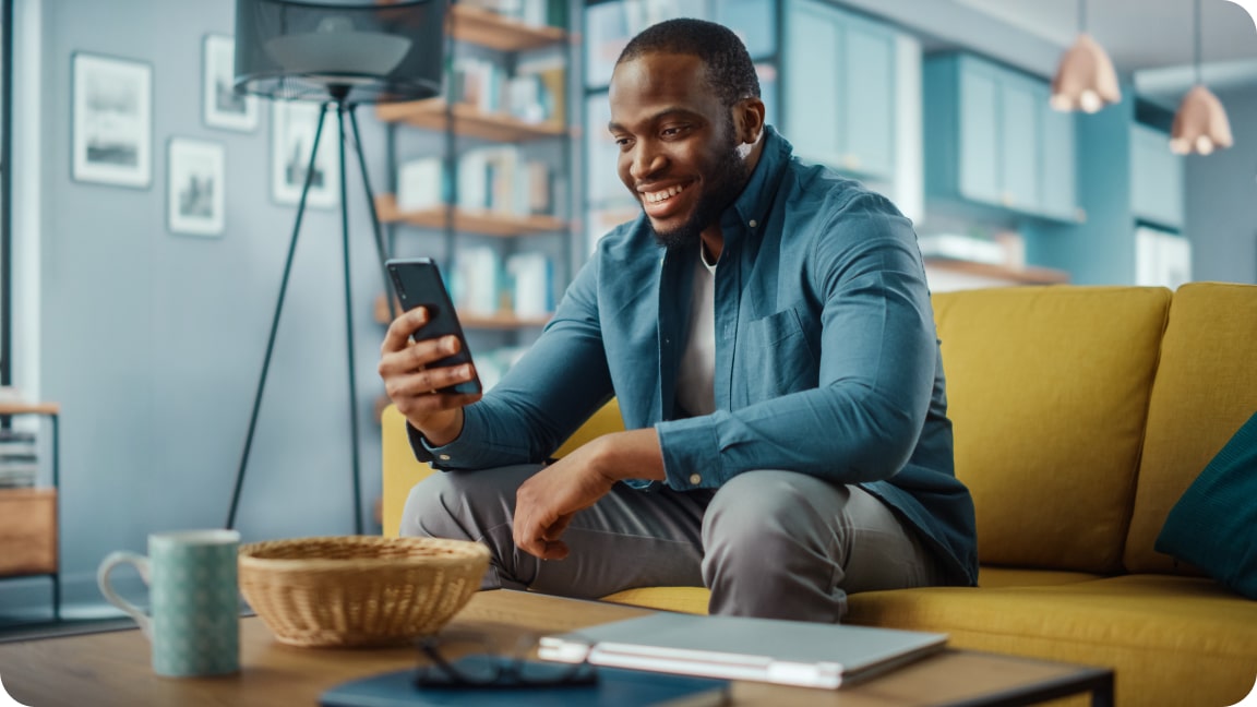 Sitting on a couch in an office area, a person looks at their phone, holding it with one hand, as the smile.