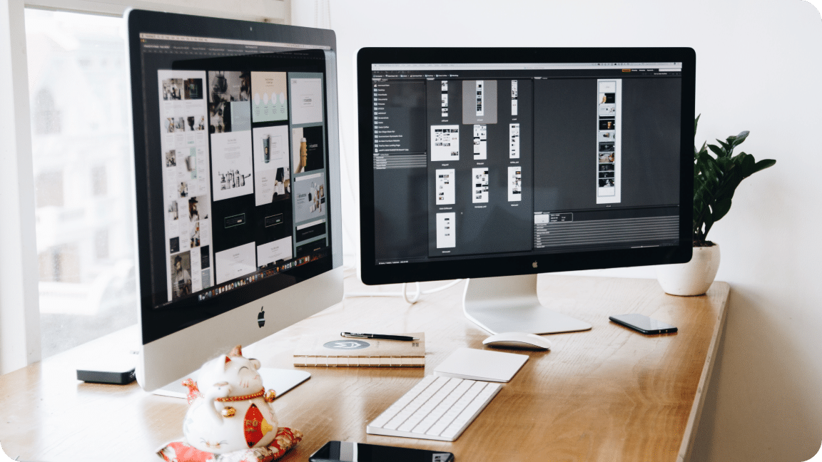 Two monitors sit on a desk displaying various design stages of a website.