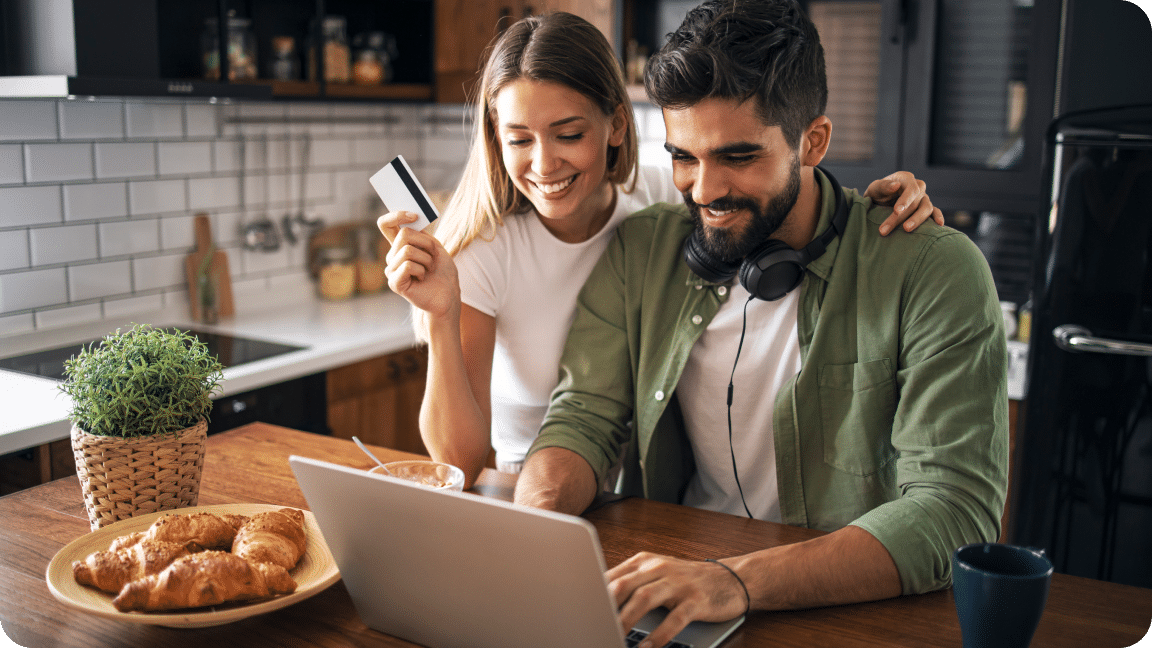 Two people at a kitchen table look at an open laptop with one holding a bank card, preparing to place an order.