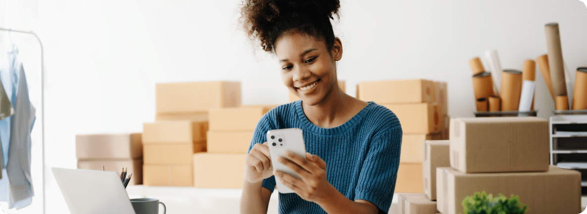 Person stands in front of shipping boxes, looking at the cellphone, smiling.