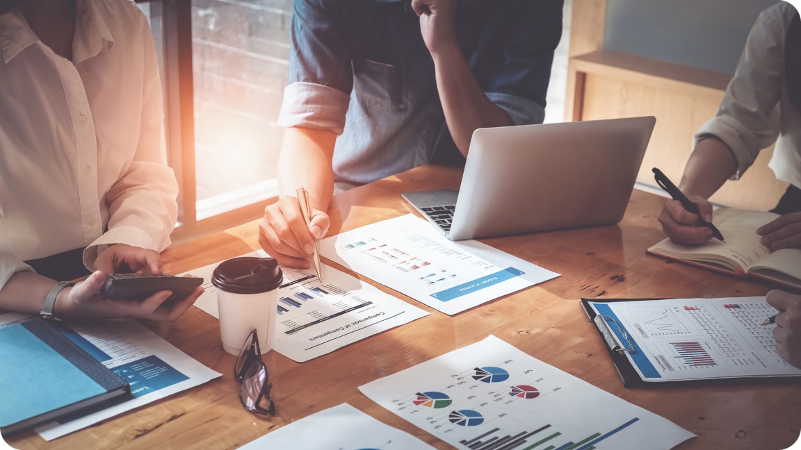 A group of people sit around a desk, charts and graphs on paper assets lay around them with notes being written.