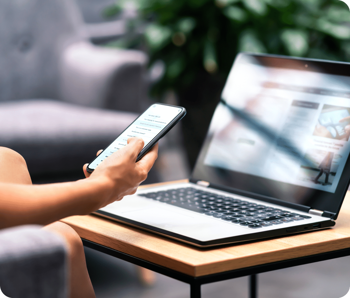 An open laptop sits on a small table with a hand holding out a cellphone.