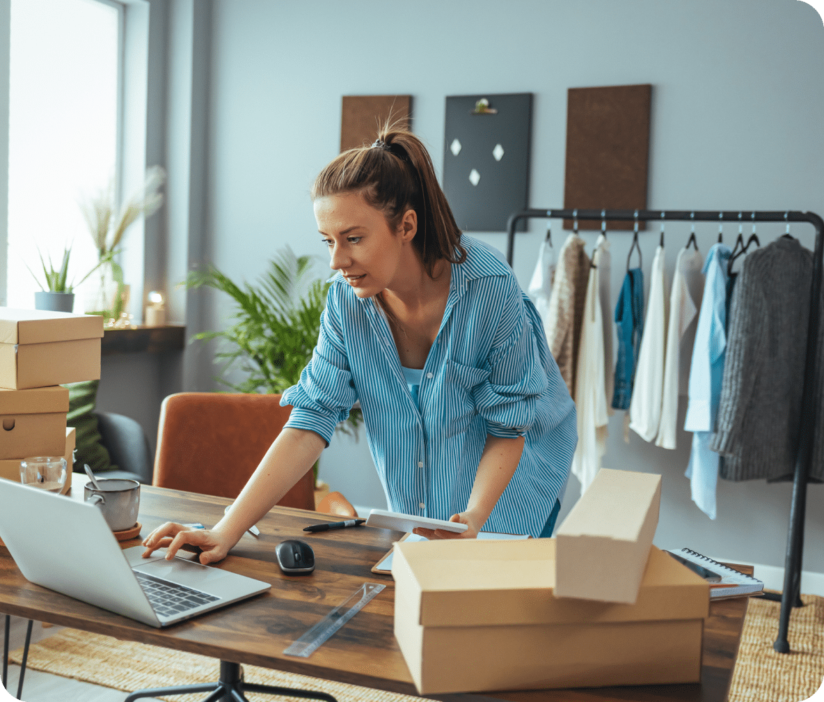Person leans over to their laptop from the front of their desk, product and shipping boxes around them.