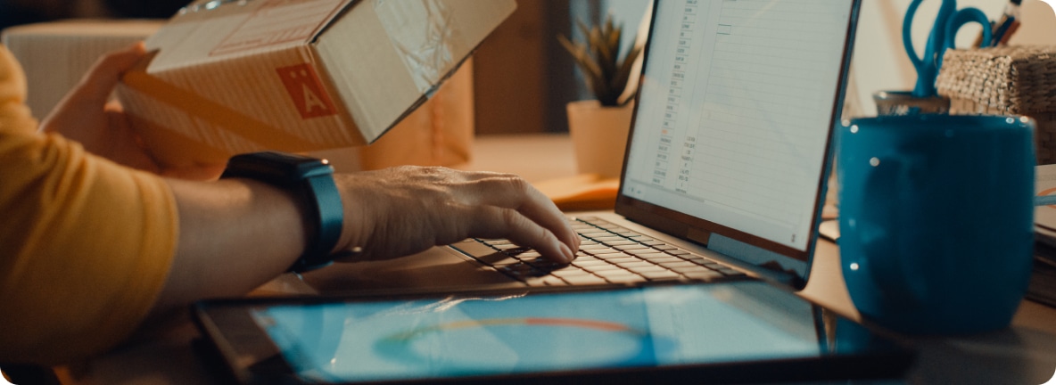 A person types on their laptop with one hand while holding a shipping box in the other.