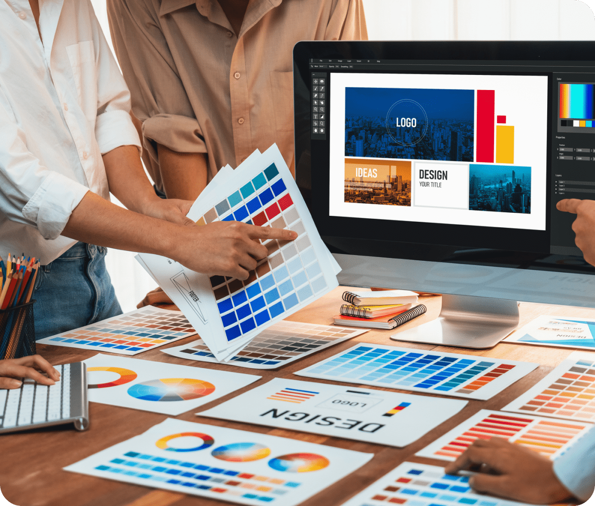 A laptop with designing software and people choosing colors from paper color charts to make assets for website advertising services.